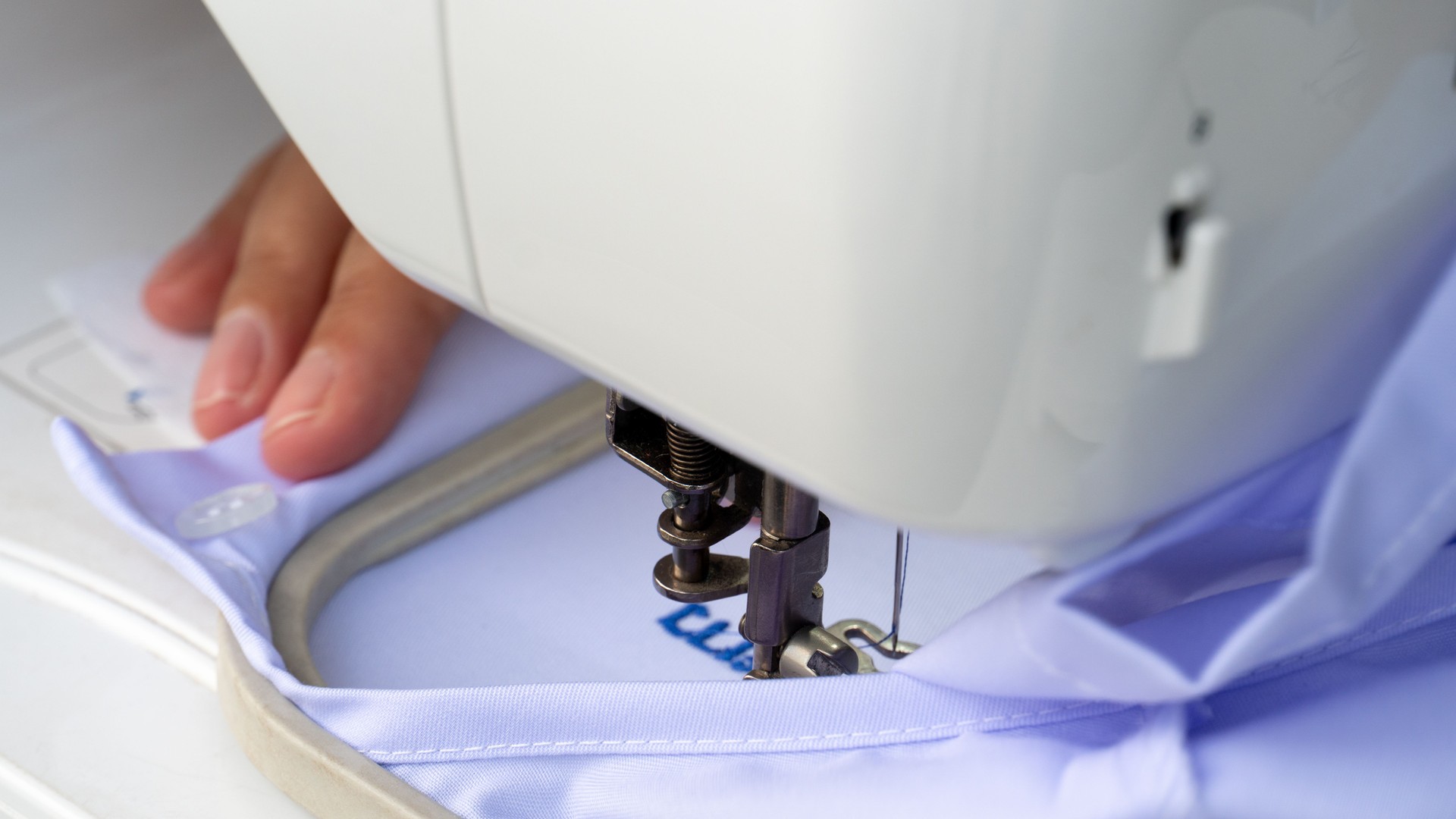 Person hand guiding light blue fabric under the needle of a white embroidery machine.